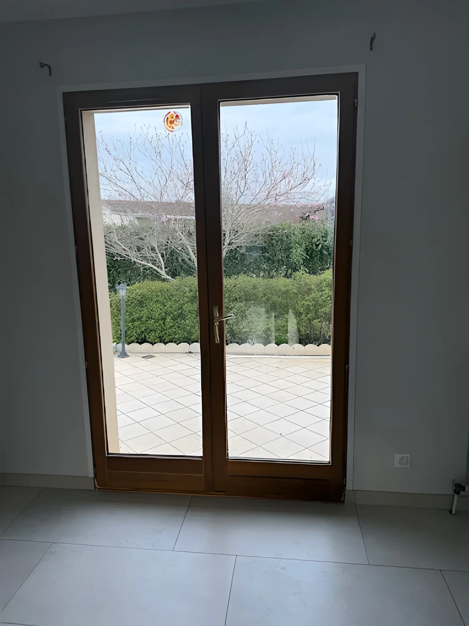 Porte-fenêtre deux vantaux ton bois posée dans une pièce au sol carrelé, terrasse et haie derrière