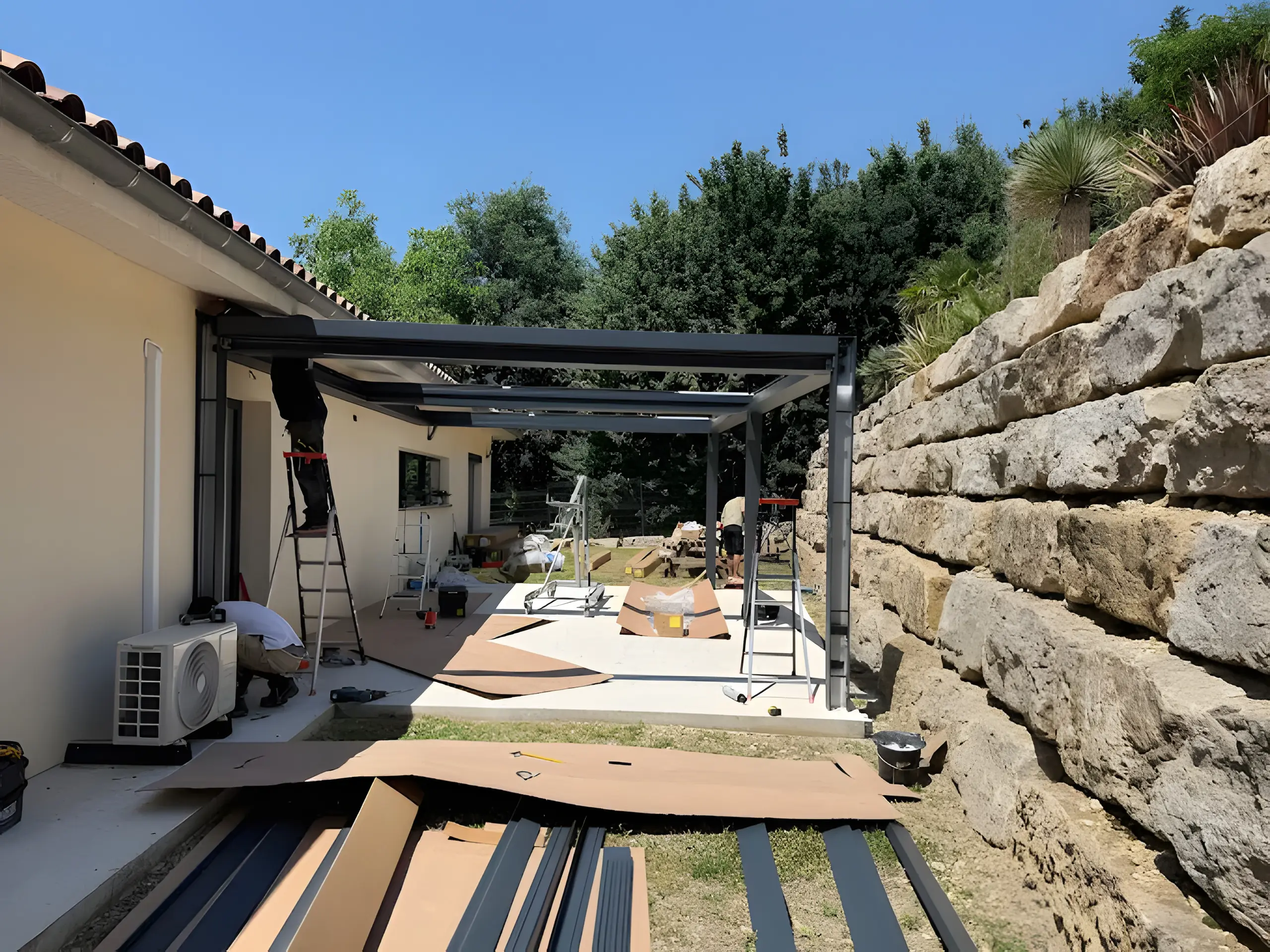 Poseurs sur escabeaux montant la structure d’une pergola aluminium anthracite contre une maison